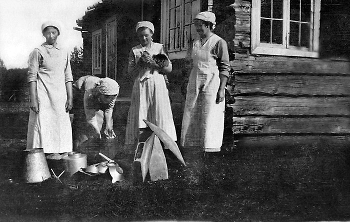 Fyra kvinnor som står utanför ett timmrat hus på marken ligger mjölkkärl och andra tillbehör. Svartvitt foto