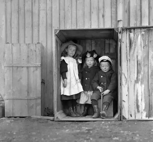 Två flickor och en pojke står i en liten låda av trä, svartvitt foto